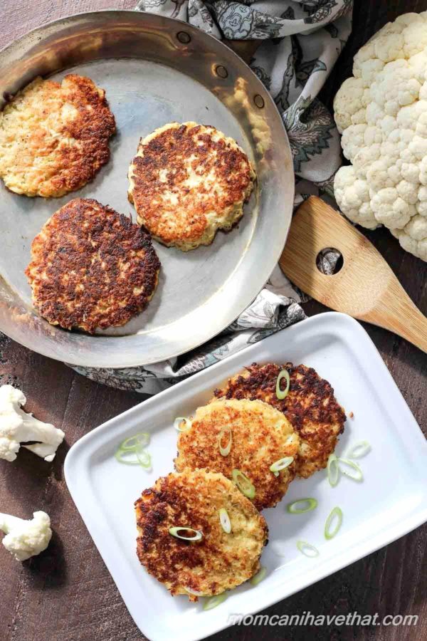 Top down photo of Cauliflower fritters or low carb hash browns stacked off-set on a white rectangular plates and in a pan with a wooden spatula to the side and pieces of raw cauliflower arranged around the plate and pan.