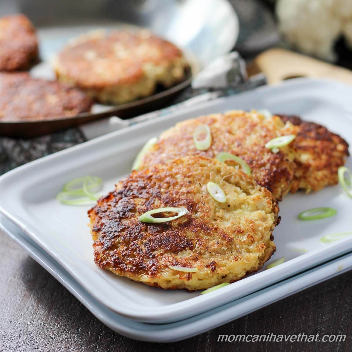 Cauliflower fritters or low carb hash browns stacked off-set on a white rectangular plates, garnished with green onions, and a skillet with more fritters behind with pieces of raw cauliflower in the background.