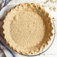 Baked low carb pie crust with fluted edge and dock marks in a pie plate on a round wire cooling rack with a blue and white kitchen towel to the left and sliced almonds around the marble surface.