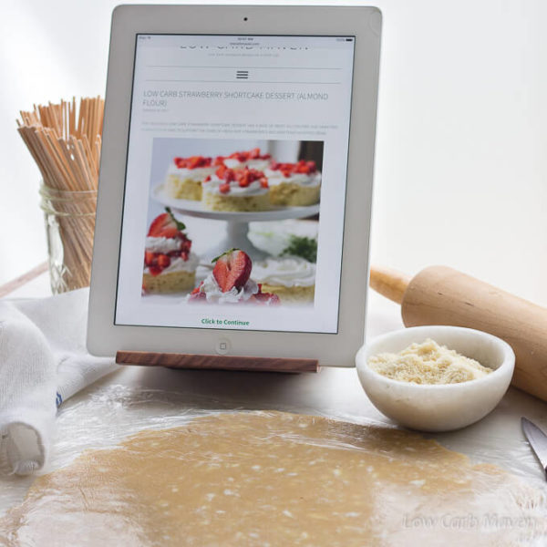 Low carb pie crust rolled out between two sheets of plastic wrap with a bowl of almond flour and rolling pin behind and an iPad in a wooden stand showing a photo of strawberry shortcakes.