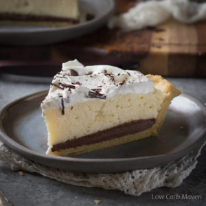 A slice of low carb black bottom pie, featuring a layer of chocolate custard and an airy chiffon layer topped with whipped cream and chocolate shavings on a dark gray plate placed on a folded white napkin with a wooden tray behind.