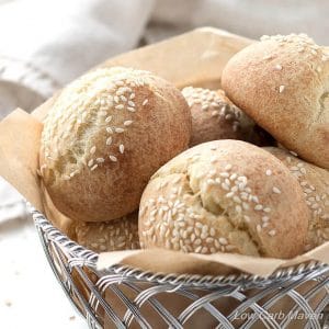 Round low carb rolls with sesame seeds and split tops in a woven aluminum basket lined with a brown piece of parchment and a beige napkin in the background.