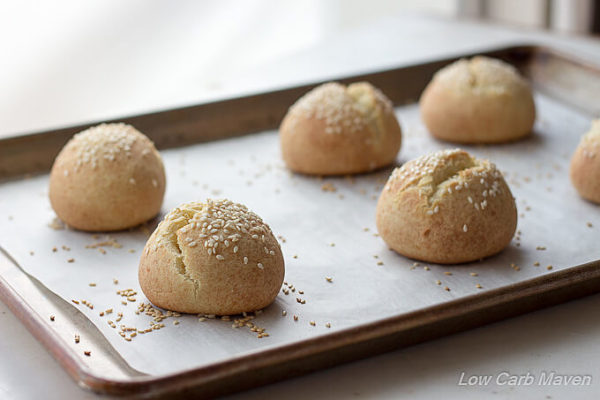 Fresh baked low carb sesame seed rolls with split tops on a parchment lined baking sheet placed on a white marble counter.