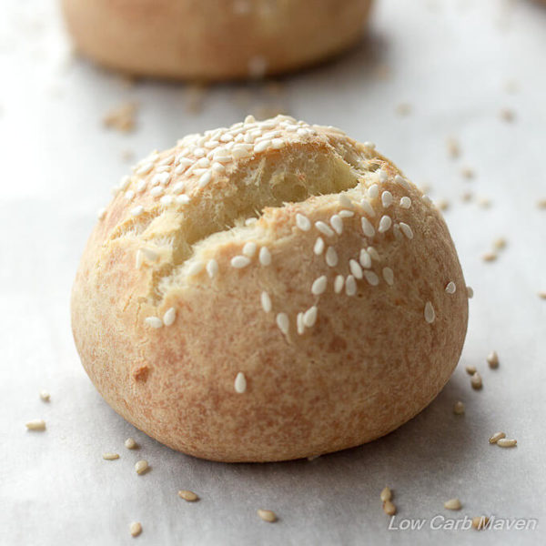 Round low carb roll with sesame seeds and a split top on a piece of parchment with another roll in the background.