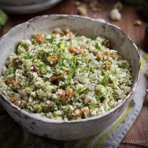 A low carb side of riced broccoli and cauliflower with walnuts in a bowl sitting on a wooden cutting board with a green striped napkin and a wooden pepper mill in the background.