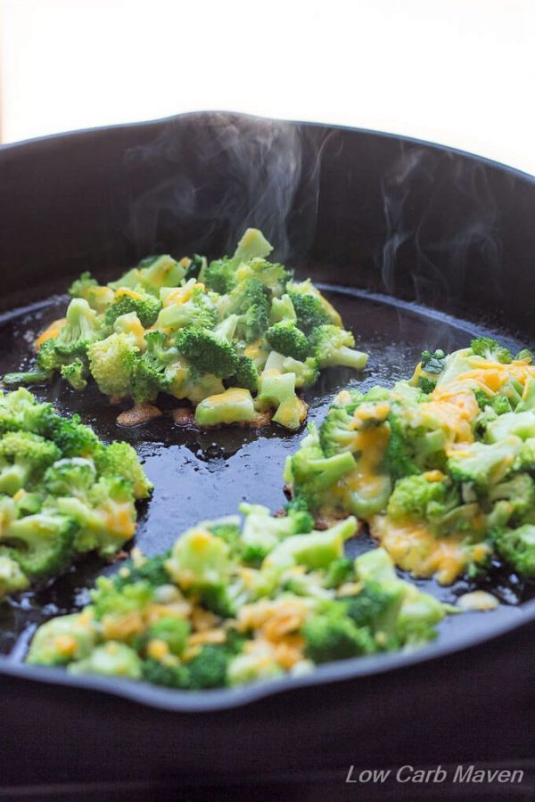 Broccoli fritters with melted cheddar cheese cooking in a cast iron pan with curls of steam rising into the air. 