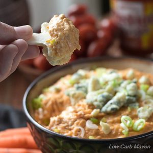 Low Carb Buffalo Chicken Dip topped with blue cheese and scallions in a brown bowl surrounded by vegetables. A piece of cauliflower is shown after being dipped into the hot appetizer.