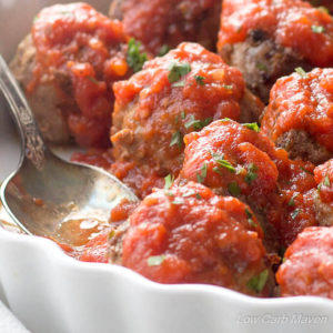 Low carb Italian meatballs in Marinara sauce and sprinkled with parsley in a white scalloped dish with a sauced spoon in a place where a meatball once sat.
