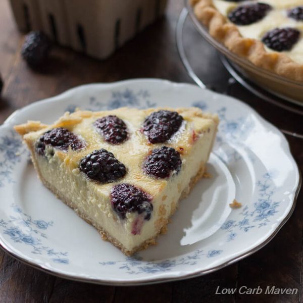 A slice of blackberry custard pie with a nice browned top and caramelized blackberries on a blue and white floral china dessert plate with a portion of the pie showing in the background to the right and a partial cardboard container of blackberries behind. 