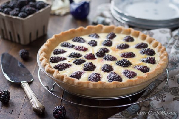 Beautifully browned custard pie with blackberries and fluted crust in a glass pie plate sitting on a round wire rack with blackberries and a silver pie server to the left, a pint of berries and china dessert plates behind and a blue and brown floral napkin to the right.