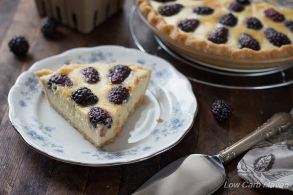 A slice of blackberry custard pie (no-bake buttermilk pie) on a blue and white floral china plate with a silver pie server to the right and the whole pie behind, with fresh blackberries on the wooden table.