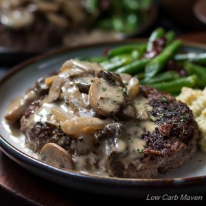 Hamburger steak and gravy of mushrooms and onions on a plate with green beans with bacon and cheesy mashed cauliflower.