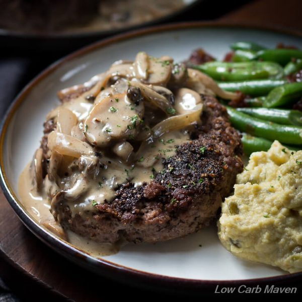 Savory looking hamburger steak with pepper crust and creamy mushroom gravy plated with mashed cauliflower and green beans with bacon.