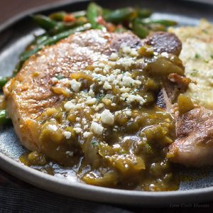 Mexican pork chops recipe with chile verde sauce and cotija cheese served with mexican green beans and tomatoes and cheesy cauliflower mash on a dark handmade plate.