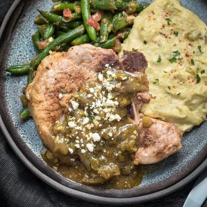 Mexican pork chops with chile verde sauce and cotija cheese served with mexican green beans and tomatoes and cheesy cauliflower mash on a dark handmade plate.