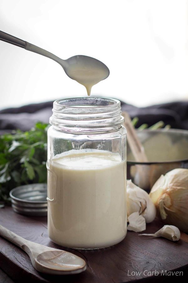 Creamy keto Alfredo sauce, Parmesan cream sauce, dripping from a spoon over a filled jar of sauce on a cutting board. A wooden spoon sits to the left of the jar with parsley, onions, garlic and a pot in the background.