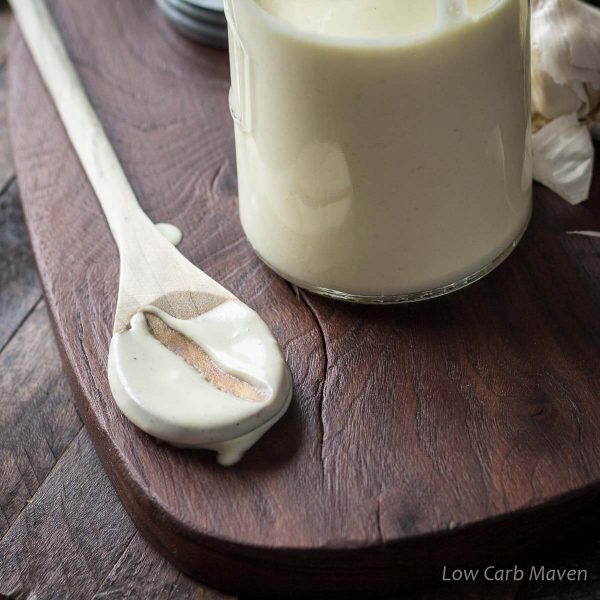 Creamy keto Alfredo sauce on a wooden spoon with a finger-track showing the thickness of the sauce.  Spoon is on a cutting board with a jar of Alfredo sauce to the right.