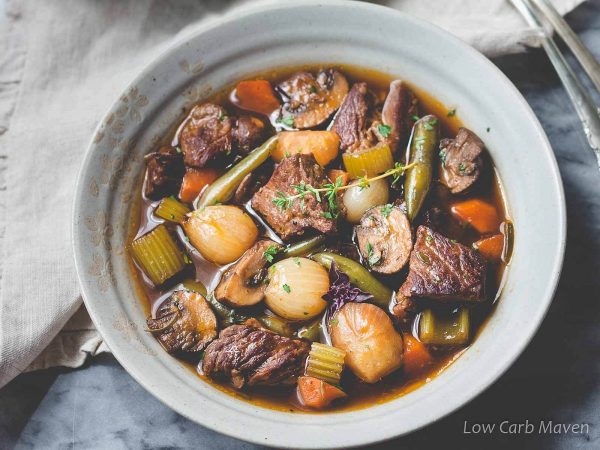 Top-down view of a bowl of hearty vegetable beef stew with carrots, celery, celery root and pearl onions in a rich beef broth and topped with a sprig of fresh thyme. 