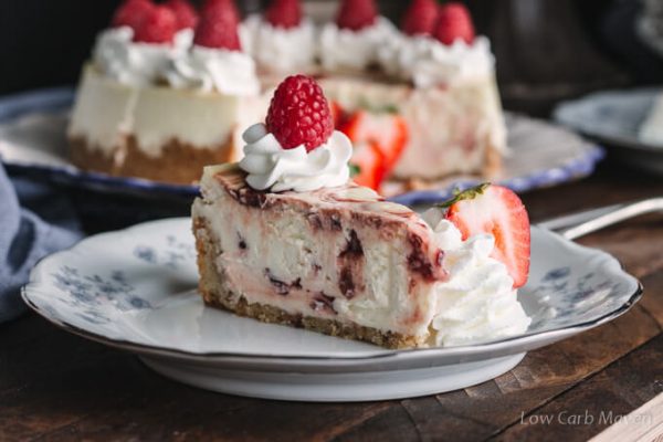 Slice of keto cheesecake with raspberry swirl on a plate with whipped cream and fresh raspberries and strawberries.