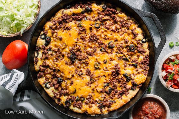 Cheesy baked keto Mexican cornbread casserole (or tamale pie) in a cast iron skillet with bowls of shredded lettuce, salsa and pico de gallo.