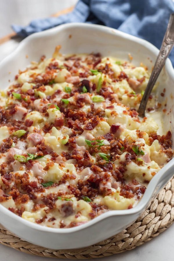 Creamy keto cauliflower mac and cheese with ham topped with bacon and sliced scallions in a white oval serving dish with serving spoon. Blue napkin in background.