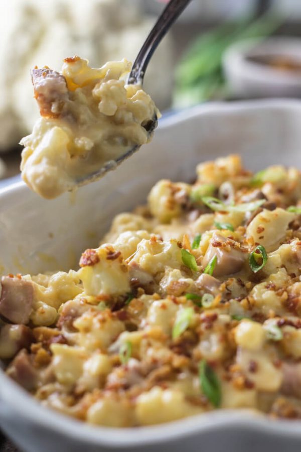 A spoon-full of creamy looking keto cauliflower mac and cheese above a white serving dish of cauliflower mac and cheese with cubed ham topped with bacon and sliced green onions.