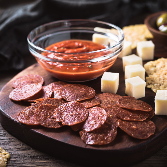 Pepperoni chips, cheese cubes, and Marinara sauce on a cutting board.