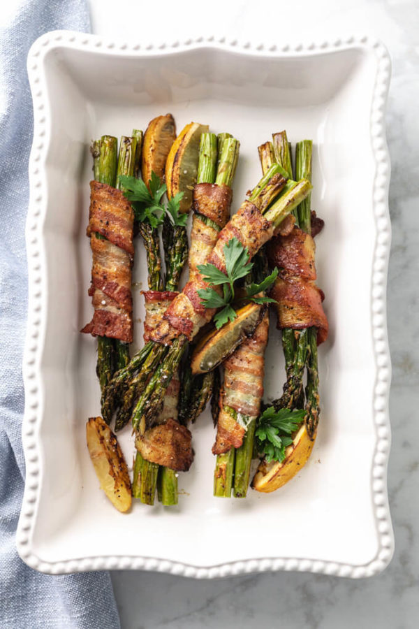 Bacon wrapped asparagus bundles with roasted lemons in a white serving dish