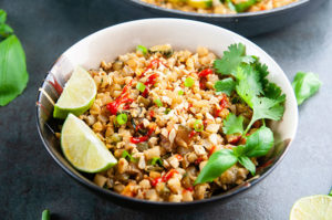 A bowl of Thai basil cauliflower fried rice on a dark background with lime wedges and cilantro as garnish
