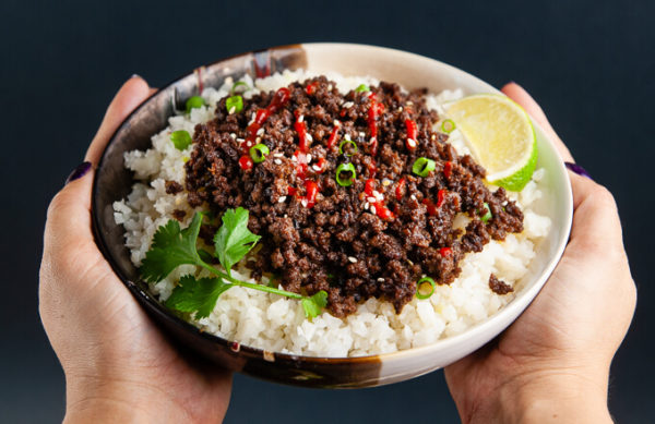 Hands holding a bowl of Korean ground beef with Siracha and lime over cauliflower rice.