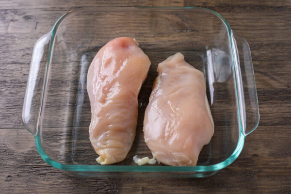 How to make oven baked chicken: two raw chicken halves in an 8x8-inch square glass baking dish on a table. 