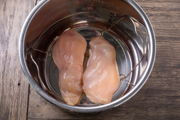 How to make shredded chicken in a pressure cooker: Two raw chicken breasts on a wire rack in an electric pressure cooker sleeve on a table.