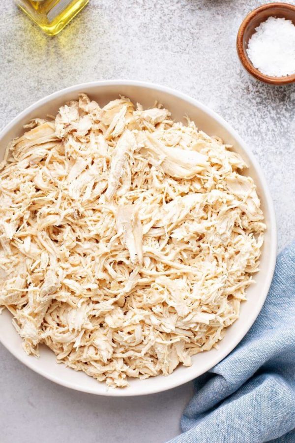 How to make shredded chicken (6 ways). Cooked shredded chicken in a bowl with blue napkin, salt and olive oil in picture.