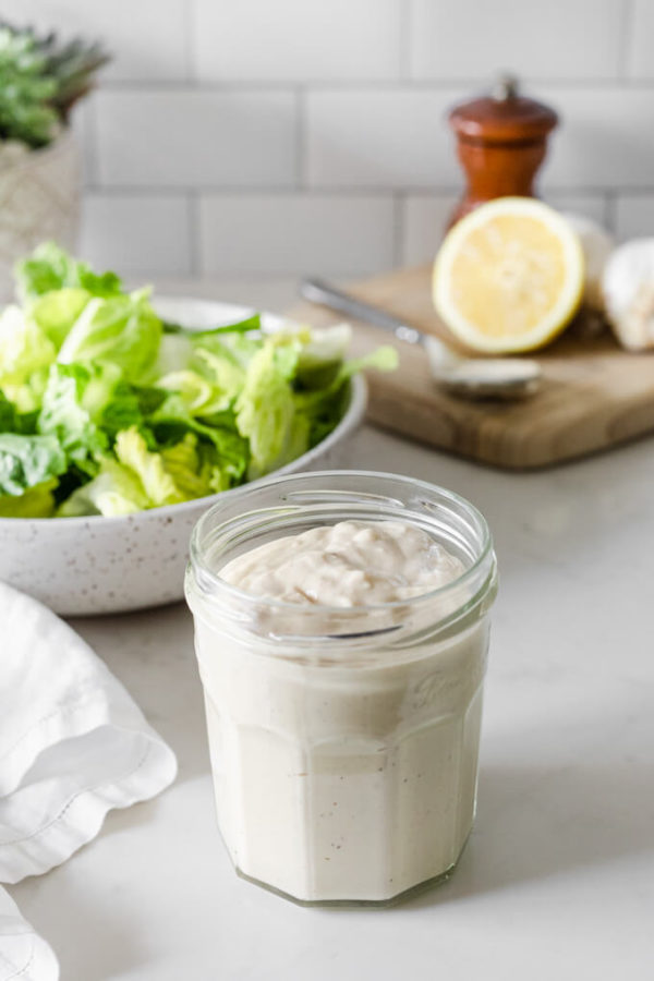 Caesar salad dressing in a glass jar with bowl of romaine lettuce and cutting board with lemons and garlic behind. 