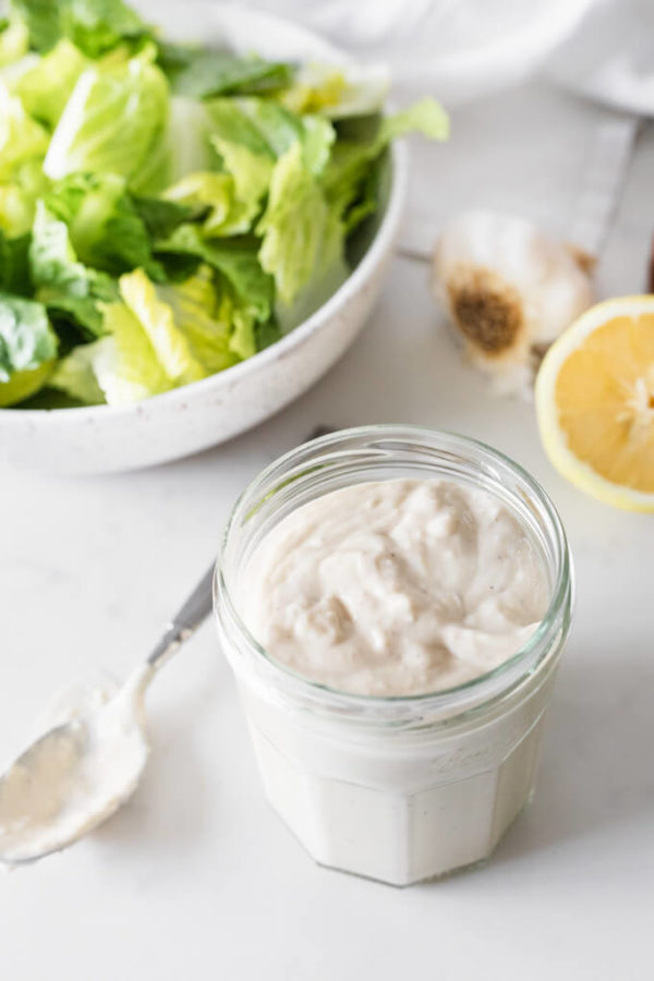 Easy Caesar salad dressing in a glass jar with spoon to side. Bowl of romaine and lemons behind.
