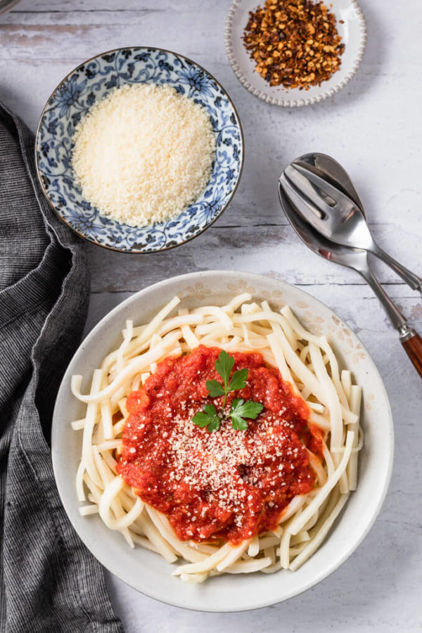 Serving of low carb pasta with keto marinara in a bowl with fork and spoon to the side and smaller bowls of grated Parmesan cheese and red pepper flakes.