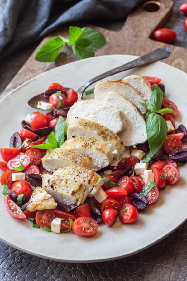 Sliced chicken breast over a Mediterranean salad with tomatoes, kalamata olives, feta cheese and fresh herbs on a platter with spoon.