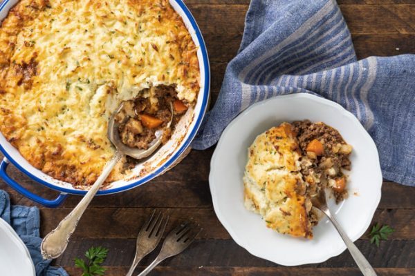 keto shepherd's pie in casserole dish with with serving spoon & individual serving in white bowl to the right with blue striped napkin.