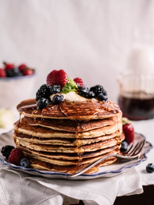 Stack of keto coconut flour pancakes with butter, mixed berries and syrup on blue edged plate with fork. Syrup and bowl of berries behind.