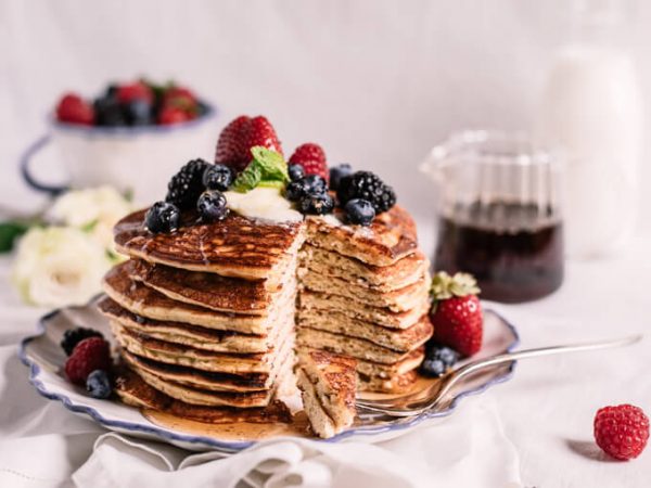 Tall stack of keto coconut flour pancakes with butter, mixed berries and syrup with missing wedge showing fluffy texture. Carafe of syrup and bowl of berries in background.