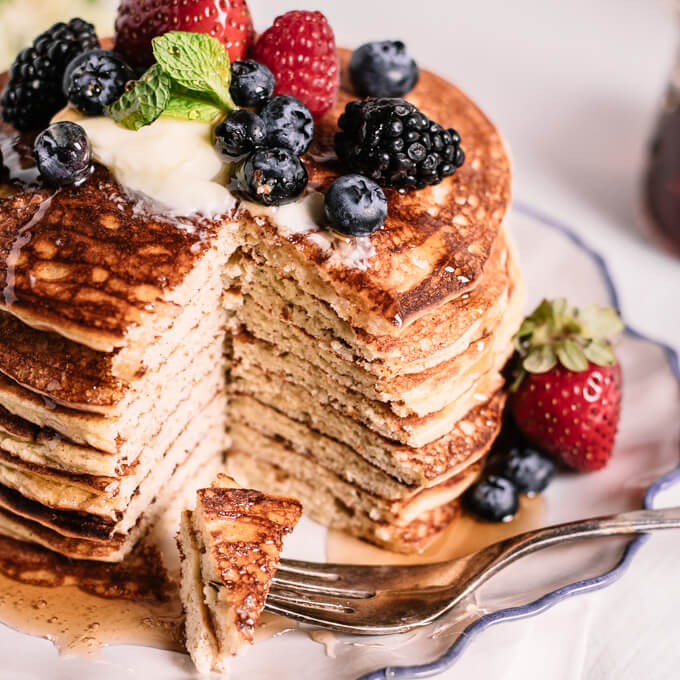 Tall stack of keto coconut flour pancakes with butter, mixed berries and syrup with fork on blue edged plate. Wedge of pancake stack removed showing layers.