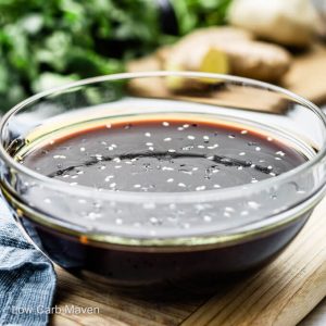 keto teriyaki sauce with sesame seeds in glass bowl with blue napkin to left and ingredients behind.