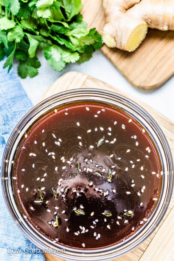 Sugar free teriyaki sauce with sesame seeds in bowl with blue napkin to left and ingredients behind.