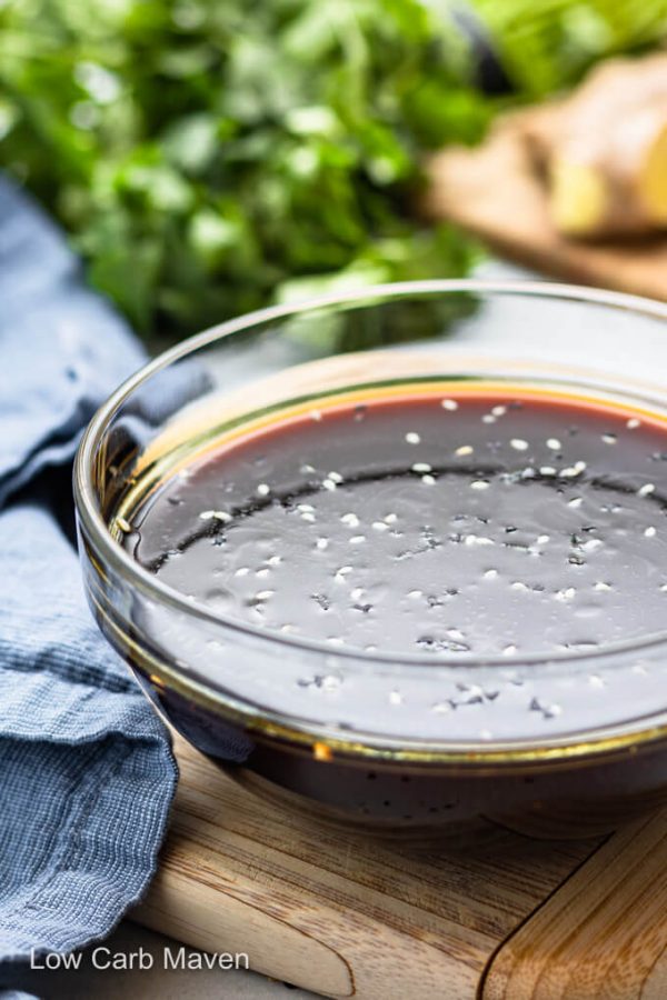 keto teriyaki sauce with sesame seeds in glass bowl with blue napkin to left and ingredients behind.