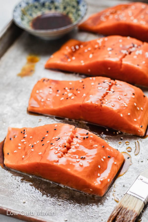 Raw salmon filets with keto teriyaki glaze and sesame seeds on sheet pan with bowl of sauce and pastry brush.