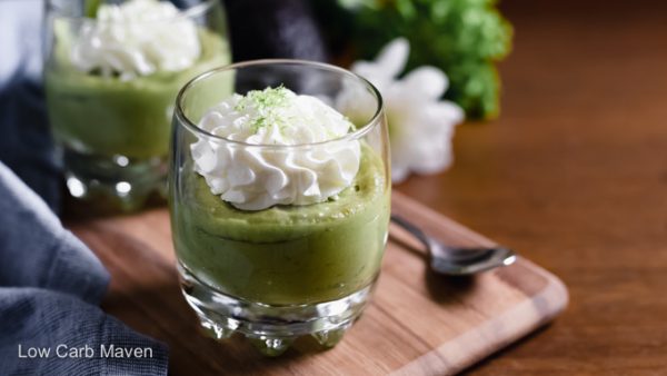 Two Keto avocado puddings in glasses with blue napkin, spoon, avocados and flowers surrounding.