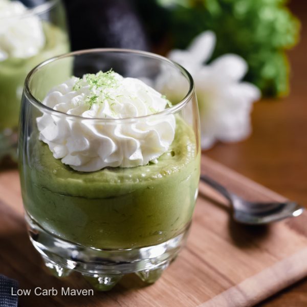 A small glass of keto avocado pudding topped with whipped cream and lime zest with spoon, avocado and white flower behind.