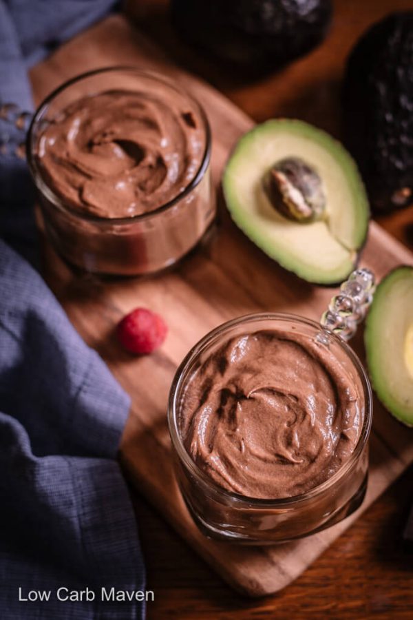 Two keto chocolate avocado puddings in cups with halved avocado on a cutting board with blue napkin to side