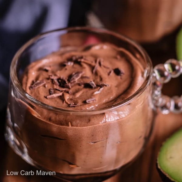 Keto chocolate avocado with shaved chocolate in a cup with avocado half and blue napkin behind.